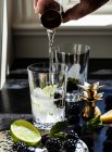 Hand pouring shot glass of vodka into cocktail glass with blackberries, lime and mint — Stock Photo