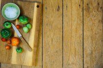 Uma tigela de sal e vários tomates com uma faca em uma tábua de corte de madeira — Fotografia de Stock