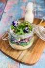 Uma salada de quinoa com cordeiros alface, radicchio, foguete, croutons, queijo de cabra e violetas com chifres em um frasco de vidro em uma placa de madeira, com molho em uma garrafa de vidro — Fotografia de Stock