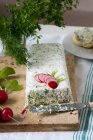 A big piece of quark cheese and herbs terrine on a marble board. Radishes, dill, parsley and a roll on a white plate in the background — Stock Photo