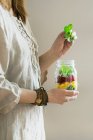 A woman in a linen dress holding a jar of fruit salad with mango, pomegranate seeds and lambs lettuce — Stock Photo