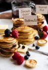 Pile di mini pancake con cartelli, con gocce di frutta e cioccolato, sciroppo e burro, su una tavola di marmo — Foto stock