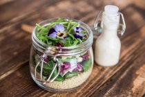 Una ensalada de quinua con lechuga de cordero, radicchio, cohete, croutons, queso de cabra y violetas con cuernos en un frasco de vidrio, con aliño en una botella de vidrio - foto de stock