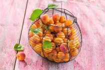 Apricots with green leaves in wire basket on pink wooden surface — Stock Photo