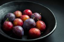 Bol de pruneaux et prunes pourpres et rouges frais dans un bol de fruits noirs sur un fond noir — Photo de stock
