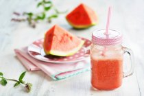 Krug mit Wassermelonen-Smoothie und Stroh vor Teller mit Melonenstücken und frischer Minze — Stockfoto