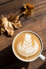 Café plano blanco capuchino con roseta o florette latte art sobre fondo de madera con hojas otoñales - foto de stock