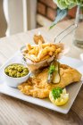 Gourmet fish and chips deep fried cod fillet in a beer batter with chips in a basket, gherkins pickle, mushy peas and half a lemon — Stock Photo
