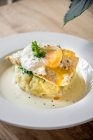 Filetto di pesce spigola su un purè di patate con tuorlo grattugiato uovo in camicia guarnito con erbe su un piatto bianco e tavolo di legno chiaro — Foto stock
