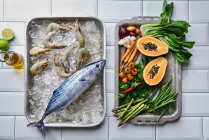 Bonito y gambas tigre sobre hielo, con verduras y papaya en bandejas de horno - foto de stock