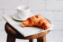 Frühstückscroissant, Tasse Kaffee und Zeitung auf einem Holzhocker — Stockfoto