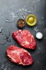Gros plan de steak de viande marbrée crue Ribeye sur fond de pierre rustique noire avec assaisonnements, huile d'olive — Photo de stock