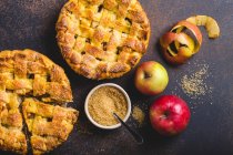 Top view of delicious homemade apple pie with cut slice, fresh apples, peel, cane sugar on brown rustic stone background — Stock Photo