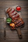 Steak de viande marbrée grillé ou frit et tranché avec fourchette, tomates comme plat d'accompagnement et différentes sauces sur planche à découper en bois, vue sur le dessus, gros plan — Photo de stock