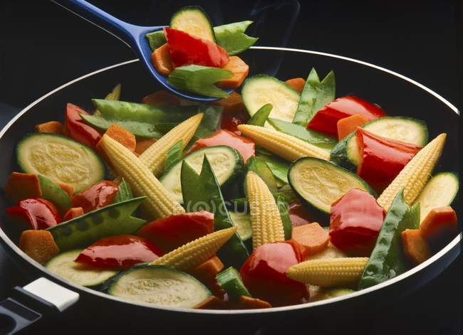 Faire revenir les légumes dans une casserole et une cuillère bleue sur fond noir — Photo de stock