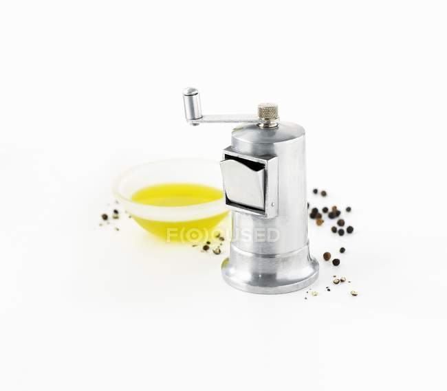 Closeup view of pepper grinder with peppercorns and oil in bowl on white surface — Stock Photo