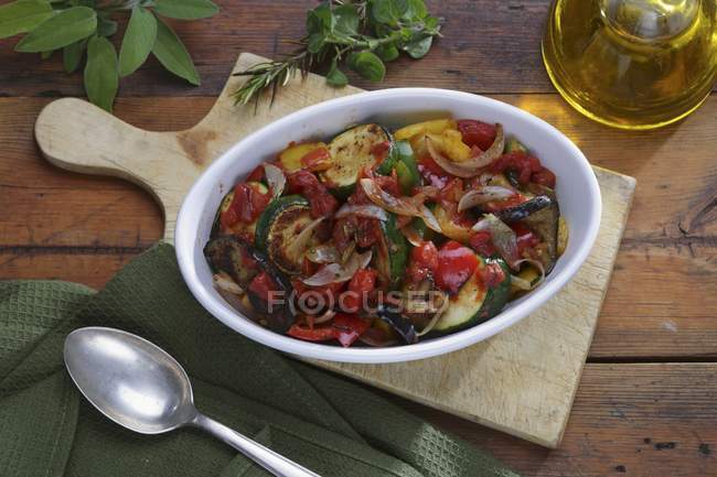 Рататуй, оливковое масло и свежие травы на белой тарелке над деревянным столом — стоковое фото