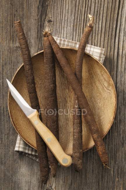 Frische Schwarzwurzeln auf einem Holzteller mit einem Messer über der weißen Oberfläche — Stockfoto