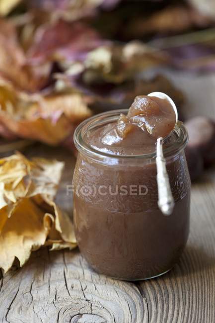 Closeup view of chestnut cream in jar on wooden surface with autumnal leaves — Stock Photo