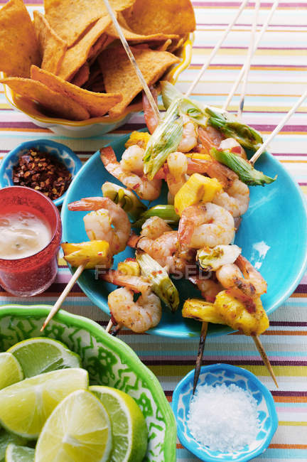 Mexican shrimp kebabs; limes; tortilla chips over colored tablecloth — Stock Photo