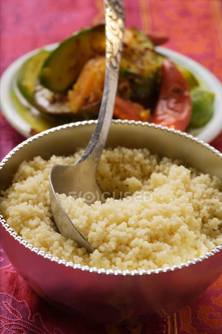 Couscous dans un bol en argent, assiette de légumes derrière sur surface rouge — Photo de stock