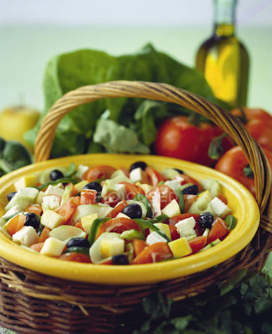 Mixed salad with cucumber, tomatoes, olives and feta on yellow plate over straw basket — Stock Photo