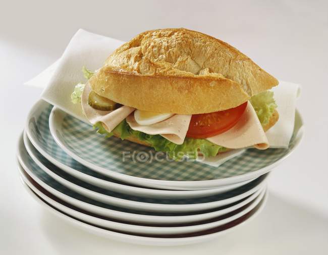 Bread roll filled with cold cuts, egg, gherkin, tomato  on stacked plates  on white surface — Stock Photo