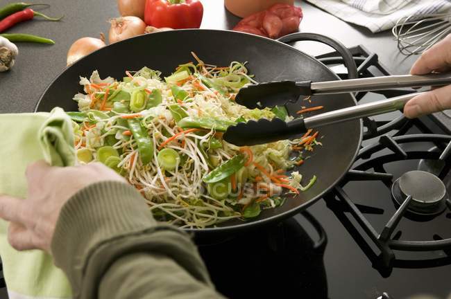 Verduras en wok siendo removidas por pinzas en las manos - foto de stock