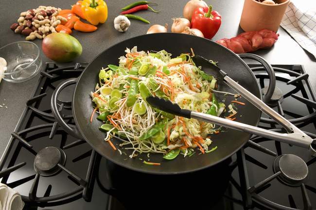 Légumes dans wok sur plaque de cuisson, ingrédients asiatiques derrière — Photo de stock