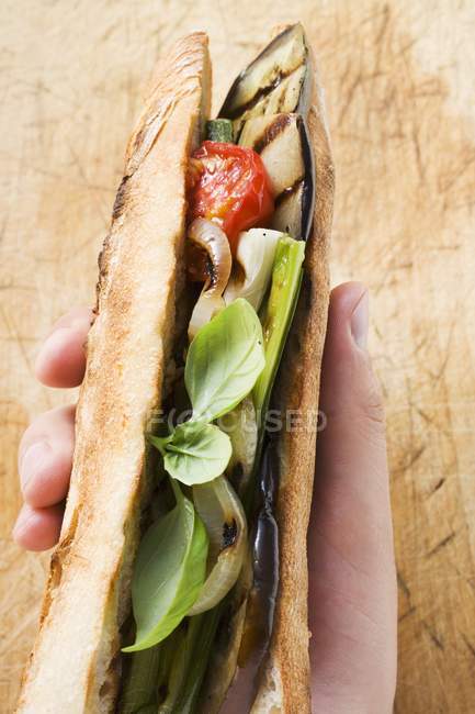 Hand holding grilled vegetables and basil in baguette over wooden surface — Stock Photo
