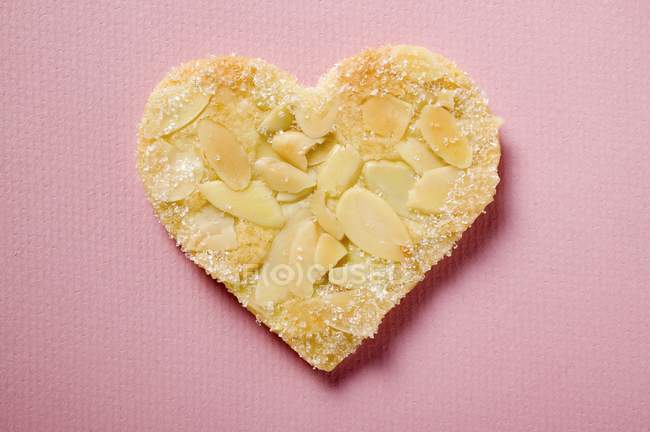 Vue rapprochée d'un cœur pâtissier avec des amandes écaillées et du sucre à la surface rose — Photo de stock