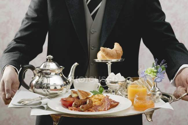 Butler serving English breakfast on tray — flowers, backdrop - Stock