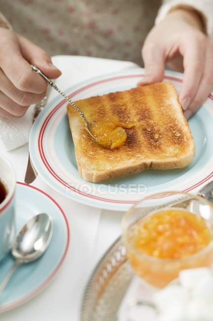 Mãos Espalhando marmelada laranja na torrada sobre placa — Fotografia de Stock