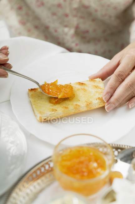 Mãos com colher Espalhando marmelada laranja na torrada na placa branca — Fotografia de Stock