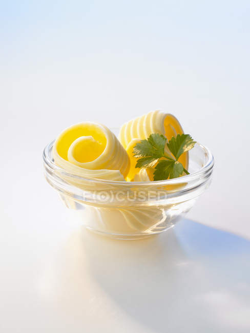 Vista close-up de cachos de manteiga em uma tigela de vidro pequeno com erva na superfície branca — Fotografia de Stock