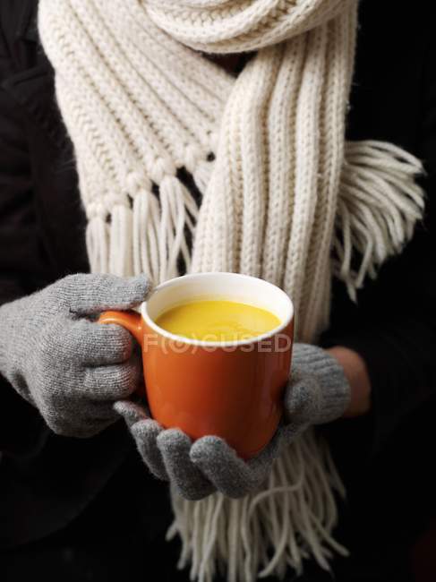 Una taza de sopa de calabaza en las manos con guantes contra la bufanda - foto de stock