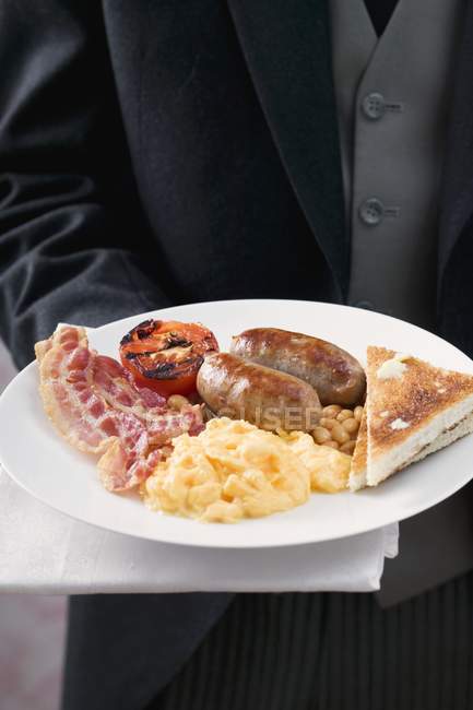 English breakfast: bacon, egg, sausage and beans, on white plate in hands, midsection — Stock Photo