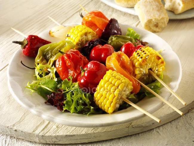 Kebab vegetali colorati su foglie di insalata su piatto bianco su scrivania di legno — Foto stock