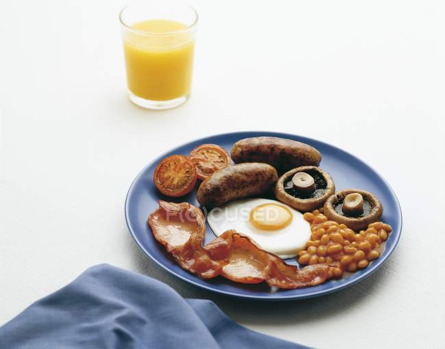 Colazione all'inglese con fagioli al forno, uova fritte, pancetta e salsiccia su piatto blu su superficie bianca — Foto stock