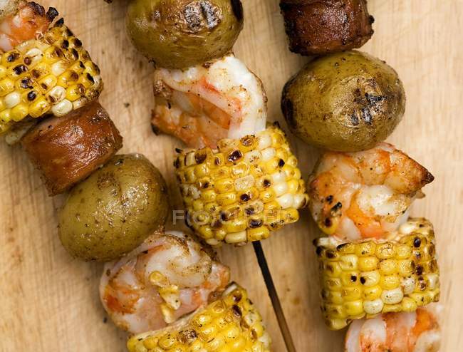 Kabobs de crevettes, maïs, saucisses et pommes de terre sur surface en bois — Photo de stock