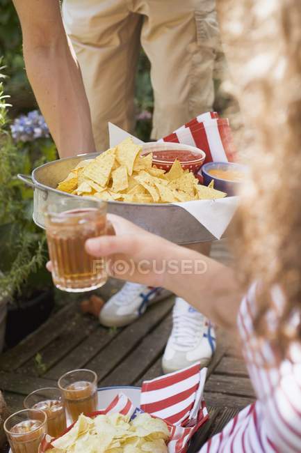Vue recadrée de personnes tenant des plateaux de chips de tortilla, trempettes et thé glacé — Photo de stock