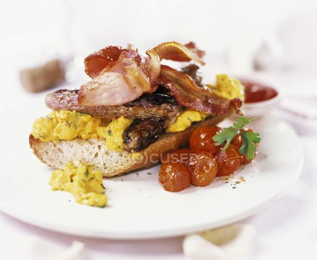 Ovo mexido, salsichas, bacon e tomates na baguete no prato branco — Fotografia de Stock
