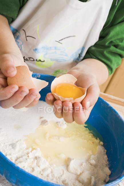 Vista de primer plano del niño añadiendo huevo a la harina y mantequilla en un tazón - foto de stock