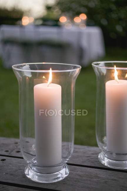 Vista de cerca de dos velas encendidas en los parabrisas de cristal en la mesa de madera del jardín - foto de stock