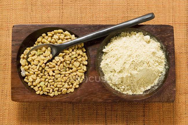 Немолотые и молотые семена пажитника с ложкой в секционированной деревянной тарелке — стоковое фото