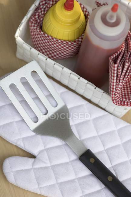 Elevated view of ketchup and mustard in basket near barbecue glove and spatula — Stock Photo
