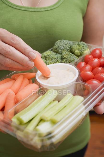 Cropped view of woman dipping carrot in sauce on plastic tray of vegetables — Stock Photo