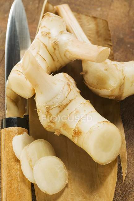 Raízes de galanga frescas e faca asiática na superfície de madeira — Fotografia de Stock