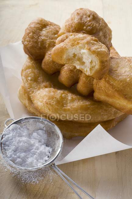Vista de cerca de pasteles fritos en papel con azúcar en polvo en el tamiz - foto de stock