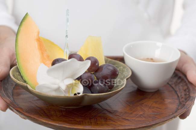 Primer plano vista recortada de la persona sosteniendo plato de fruta y tazón de té en bandeja - foto de stock